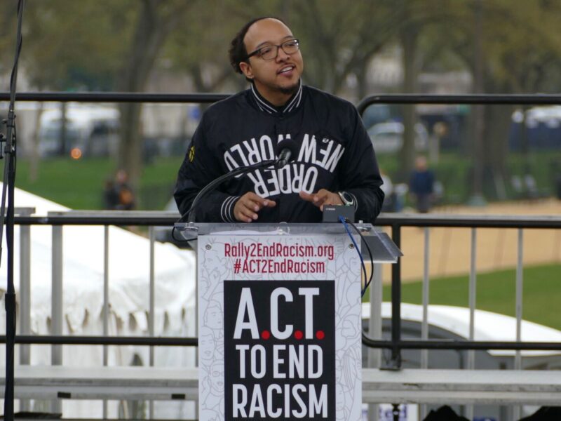 a man in a jacket addresses a rally behind a podium reading "ACT to end racism"