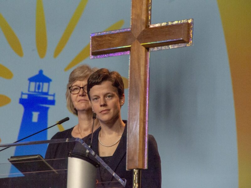 a person stands on stage behind a microphone and large, wooden cross