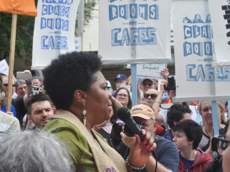 a woman holding a microphone addresses a crowd holding signs that say, "Class rooms not cages"