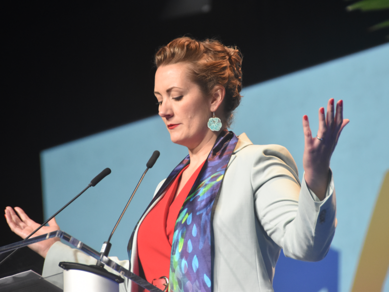 a pastor stands behind a podium with her arms stretched out and head bowed